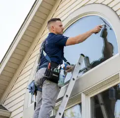 Professional window cleaner on a ladder using a squeegee to clean an exterior arched window, Oakland, CA.